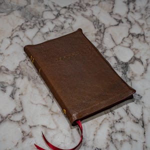 May include: A brown leather-bound book with gold lettering on the cover and spine. A red ribbon bookmark extends from the book. The book is resting on a marble surface.