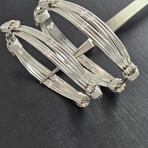 May include: Two silver-toned bangles, each constructed from multiple thin, parallel wire strands. The strands are gathered and secured at two points on each bangle with tightly coiled silver-toned wire wraps, creating a structured, multi-layered appearance.