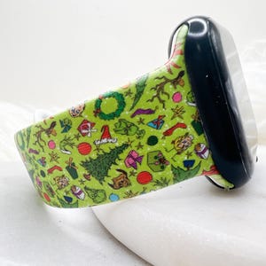 May include: A lime green silicone watch band featuring a repeating holiday-themed pattern of Christmas trees, wreaths, wrapped gifts, stockings, and small green characters. The band is attached to a black rectangular watch face housing.