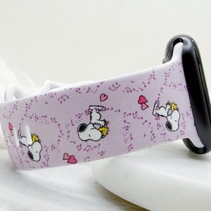 Puede incluir: Correa de Apple Watch de color lila claro con ilustraciones de Snoopy, notas musicales y corazones rosas. La correa tiene un interior blanco y un conector de reloj negro. El diseño es juguetón y caprichoso.