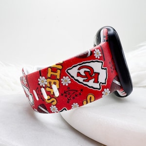 May include: A red silicone watch band featuring a repeating pattern of white daisies, brown footballs, and the Kansas City Chiefs logo in white and red. The band includes yellow text and black tactical play diagrams on a vibrant red background.