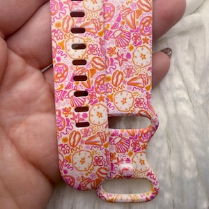 May include: A silicone watch band featuring a repeating pattern of pink, orange, and white seashells, starfish, and small floral shapes. The band includes a series of rectangular adjustment holes and a secure loop fastener for a watch attachment.