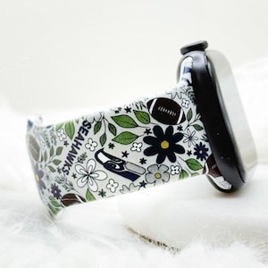 May include: A black smartwatch with a white silicone band featuring a repeating pattern of green leaves, dark blue flowers, brown footballs, and the Seattle Seahawks logo with the text SEAHAWKS printed vertically along the side of the watch strap.