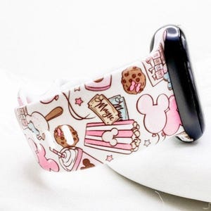 May include: A white silicone watch band featuring a repeating pattern of pink mouse-shaped balloons, brown chocolate chip cookies, pink and white striped popcorn containers, and a rectangular ticket with the word Magic printed in black script.
