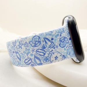 May include: A white silicone watch band featuring a repeating pattern of blue hand-drawn illustrations of various seashells, starfish, and sand dollars, attached to a black watch connector piece on one side of the strap.