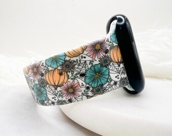 Könnte beinhalten: Ein Uhrenarmband aus Silikon mit weißem Hintergrund und einem sich wiederholenden Muster aus orangefarbenen Kürbissen, türkisfarbenen und rosafarbenen Blumen, kleinen weißen Geistern sowie schwarzen Spinnennetzen mit winzigen Spinnen.