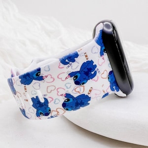 Könnte beinhalten: Ein weißes Silikon-Uhrenarmband mit einem sich wiederholenden Muster aus blauen Stitch-Figuren und kleinen rosa sowie orangefarbenen Herzumrissen. Das Armband ist an einem schwarzen Uhrenanschluss befestigt und zeigt ein flexibles Design.