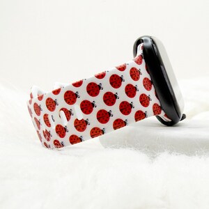 May include: A white silicone watch band featuring a repeating pattern of small red ladybugs with black spots. The band is attached to a black watch casing and includes multiple oval-shaped adjustment holes along the length of the strap.