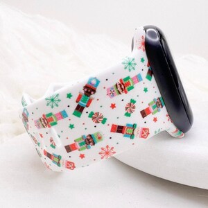 Könnte beinhalten: Ein weißes Silikon-Uhrenarmband mit einem wiederkehrenden Muster aus bunten Nussknacker-Figuren, Pfefferminzbonbons sowie grünen und roten Schneeflocken. Das Armband ist an einer Seite mit einem schwarzen Uhrenanschlussstück versehen.