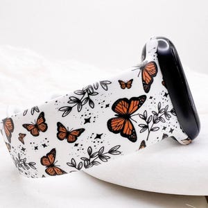 May include: A white silicone watch band featuring a repeating pattern of orange and black monarch butterflies, black line-art foliage, and small black star accents. The band includes a black connector piece attached to one end of the strap.
