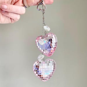May include: A silver keychain with a heart-shaped clasp holding a chain with two pink disco ball hearts. The hearts are made of small, reflective squares and are adorned with clear, teardrop-shaped beads.