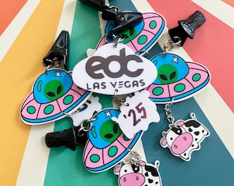 May include: A set of five colorful keychains featuring a pink and blue UFO design, a white and green alien design, a white and silver '25' design, and two pink and black cow designs. The keychains are attached to black clips and silver chains. The keychains also feature a white oval tag with the text 'edc LAS VEGAS'.