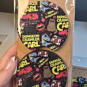May include: Two circular car coasters featuring a black background with a colorful Dungeon Crawler Carl pattern. The design includes text, a cat with sunglasses, a blue dinosaur, and red lips. The coasters are packaged in a clear plastic sleeve with a brown cardboard backing labeled Car Coaster.