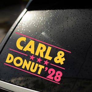 May include: A vinyl decal applied to a car window featuring the yellow text CARL & DONUT '28 with three pink stars between the lines of text. The decal is framed by two horizontal pink lines on a dark window surface covered in small water droplets.