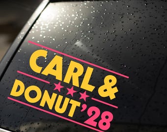Puede incluir: Un adhesivo de vinilo aplicado en la ventana de un coche con el texto amarillo CARL & DONUT '28 y tres estrellas rosas entre las líneas de texto. El adhesivo está enmarcado por dos líneas rosas horizontales sobre un cristal oscuro con gotas.