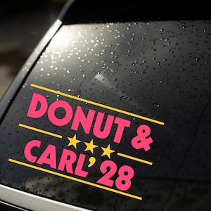 May include: A pink vinyl decal applied to a dark car window with water droplets. The decal features the text DONUT & CARL '28 in a bold sans-serif font, accented by three yellow stars and horizontal yellow lines above and below the text.
