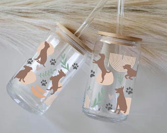 May include: Two clear glass tumblers with bamboo lids and straws. Both tumblers feature a brown dog silhouette design with paw prints, hearts, and abstract shapes in shades of brown, peach, and green.