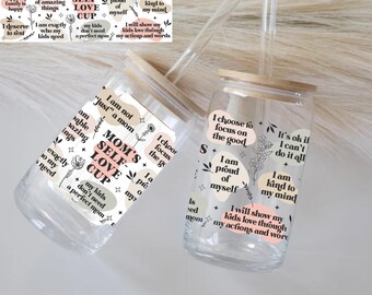 May include: Two clear glass cups with bamboo lids and glass straws. Each cup has a sticker with inspirational phrases like "Mom's Self-Love Cup" and "I am proud of myself." The cups are decorated with floral designs.