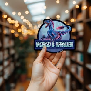 May include: A die-cut sticker featuring a blue and pink feathered dinosaur inside a circular frame, positioned above a dark blue rectangular banner with the text "MONGO IS APPALED" in light blue, sans-serif capital letters.