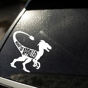May include: A white vinyl decal applied to a dark car window, featuring the silhouette of a raptor dinosaur. The text "MONGO IS APPALLED" is integrated into the body of the dinosaur, with the words following the curve of the long, thin tail.