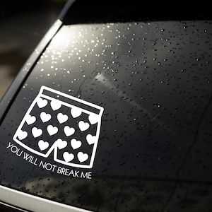 Könnte beinhalten: Ein weißer Vinyl-Aufkleber auf einer dunklen Autoscheibe, der eine Boxershorts mit Herzmuster zeigt. Unter der Boxershorts steht der Text "YOU WILL NOT BREAK ME" in einer klaren, serifenlosen weißen Schrift in Großbuchstaben.