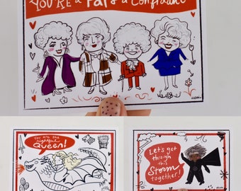 Op de afbeelding: Drie geïllustreerde kaarten met rode randen. De eerste kaart toont vier vrouwen met wit haar en rode harten. De tekst op de kaart luidt "YOU'RE a Pal & a Confidant". De tweede kaart toont een draak waarop een vrouw rijdt. De tekst op de kaart luidt "You are the rightful Queen!". De derde kaart toont een vrouw met zwart haar en een zwarte cape. De tekst op de kaart luidt "Let's get through this Storm together!".