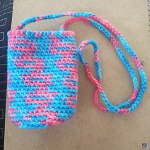 May include: A handmade crochet phone case in a vibrant mix of pink and blue. The case has a long, braided strap for wearing around the neck or shoulder. The stitch pattern creates a textured, speckled appearance.
