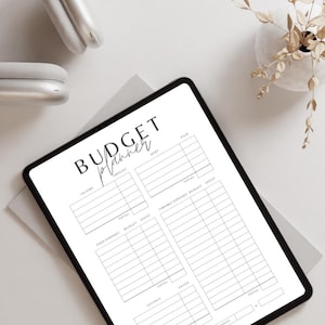 May include: A digital budget planner on a tablet with sections for income, fixed expenses, variable expenses, savings, and totals. The tablet is on a white surface with a pair of headphones and a small plant in a white pot.