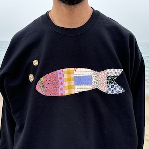 May include: Black sweatshirt featuring a colourful patchwork fish design on the front.