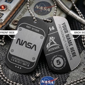 May include: Two black and silver NASA dog tags on a silver chain. One tag has the NASA logo and a reference number. The other tag says "YOUR NAME HERE" and "ASTRONAUT CANDIDATE" with the Artemis logo.