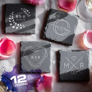 May include: A selection of square slate coasters featuring white engraved designs. The designs include initials, dates, a diamond ring, a geometric heart, and a floral wreath. The coasters are displayed on a white surface with pink flower petals.