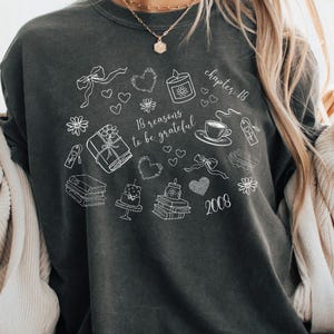 Puede incluir: Camiseta Comfort Colors gris oscuro con un diseño gráfico blanco que presenta corazones, regalos y el texto "18 razones para estar agradecido" y "capítulo 18 2008". La camiseta es de corte holgado.