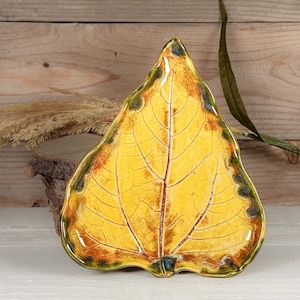 May include: A ceramic dish shaped like a large leaf with a glossy finish. The surface features a vibrant yellow center that transitions into deep orange and dark green edges, with a prominent central vein and fine crackle texture throughout.