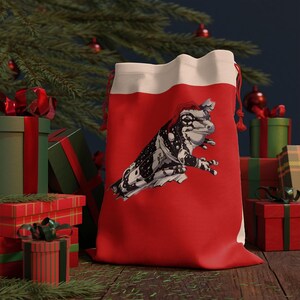 May include: A red Christmas gift bag with a white top and a drawstring closure. The bag features a black and white illustration of a frog wearing a Santa hat. Presents and a Christmas tree are in the background.