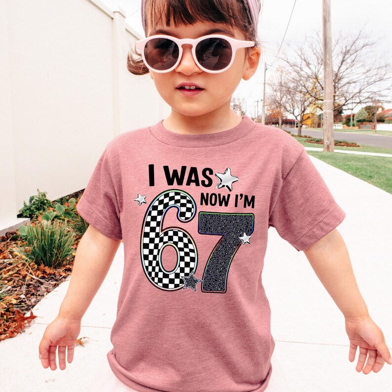 A young girl wearing a pink t-shirt with a graphic design and text, standing on a sidewalk.