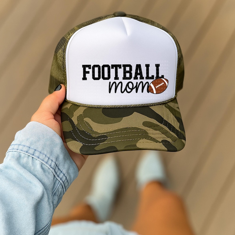 A person is holding a camouflage baseball cap with the text "FOOTBALL mom" printed on it.