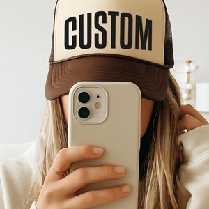 A person is taking a selfie with a brown and white hat that says "CUSTOM" on it, holding a white iPhone in front of their face.