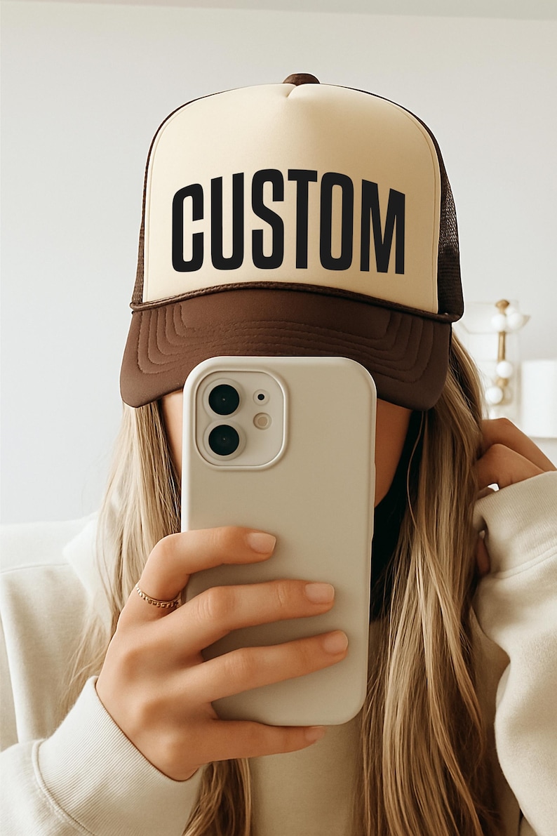 A person is taking a selfie with a brown and white hat that says "CUSTOM" on it, holding a white iPhone in front of their face.