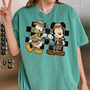 A person is wearing a green t-shirt with a cartoon image of Mickey Mouse and Donald Duck, who are depicted as adventurers or tourists.