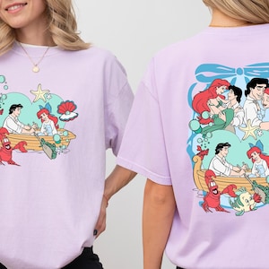 A woman wearing a pink t-shirt with a colorful graphic of characters from the Disney animated film "The Little Mermaid".