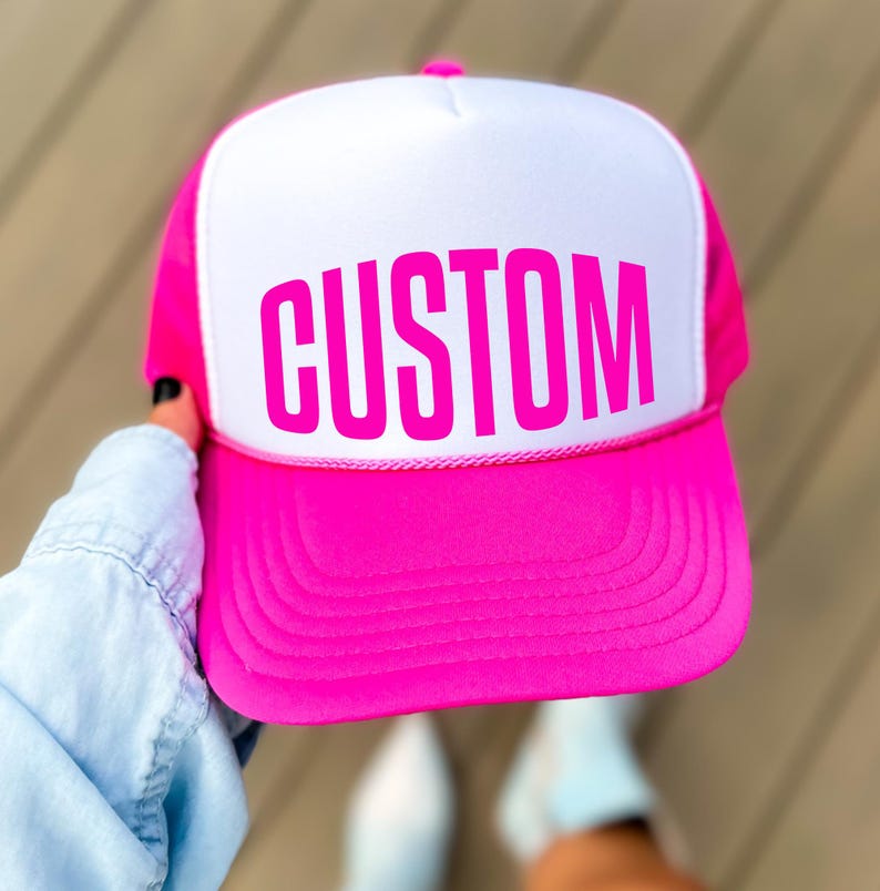 A person is holding a pink and white baseball cap with the word "CUSTOM" printed on it.