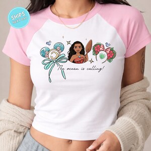 A woman wearing a white and pink t-shirt with a graphic design featuring a cartoon character and the text "The ocean is calling!"