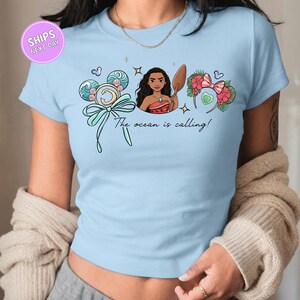 A woman wearing a light blue t-shirt with a graphic design featuring a cartoon character and text that says "The ocean is calling!"