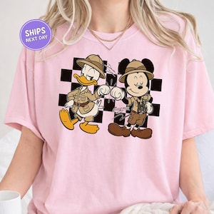 A pink t-shirt with a cartoon image of Mickey Mouse and Donald Duck, dressed in safari attire, standing next to each other.