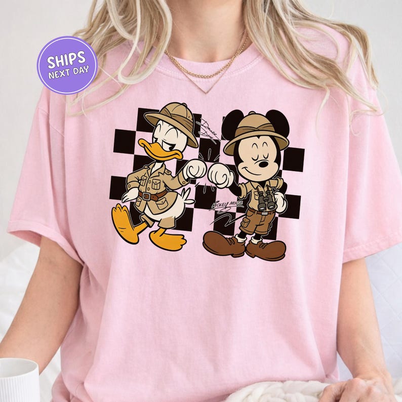 A pink t-shirt with a cartoon image of Mickey Mouse and Donald Duck, dressed in safari attire, standing next to each other.