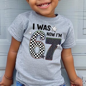 A young child wearing a gray t-shirt with the text "I WAS NOW I'M 67" printed on it.