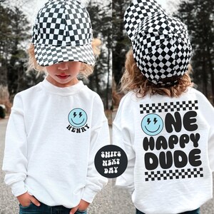 A young child wearing a white sweatshirt with a black and white checkered hat and a matching hat, along with a white sweatshirt with a black and white checkered pattern and the text "ONE HAPPY DUDE" printed on it.