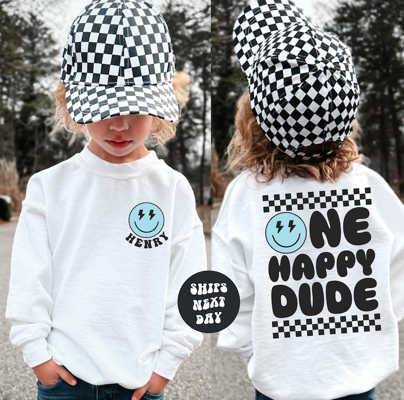 A young child wearing a white sweatshirt with a black and white checkered hat and a matching hat, along with a white sweatshirt with a black and white checkered pattern and the text "ONE HAPPY DUDE" printed on it.