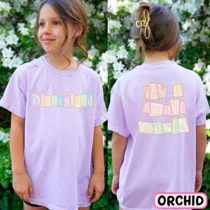 A young girl wearing a purple t-shirt with the text "Disneyland it's a small world" printed on it, standing in front of a background of white flowers.
