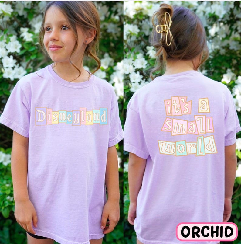 A young girl wearing a purple t-shirt with the text "Disneyland it's a small world" printed on it, standing in front of a background of white flowers.